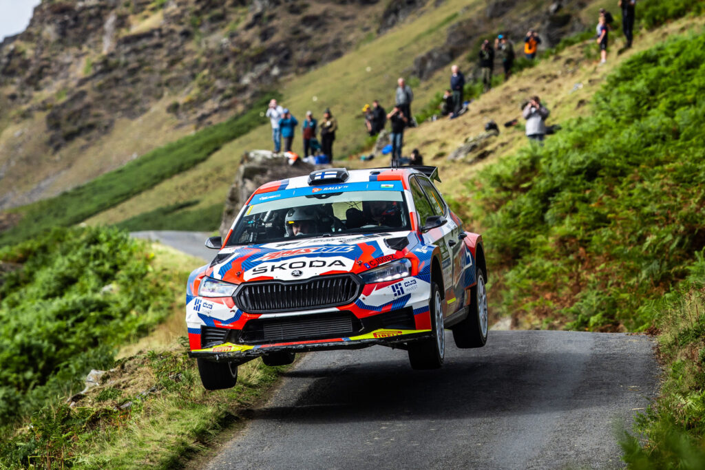 Skoda Fabia rally car jumping mid-air on a narrow mountain road with spectators in the background.