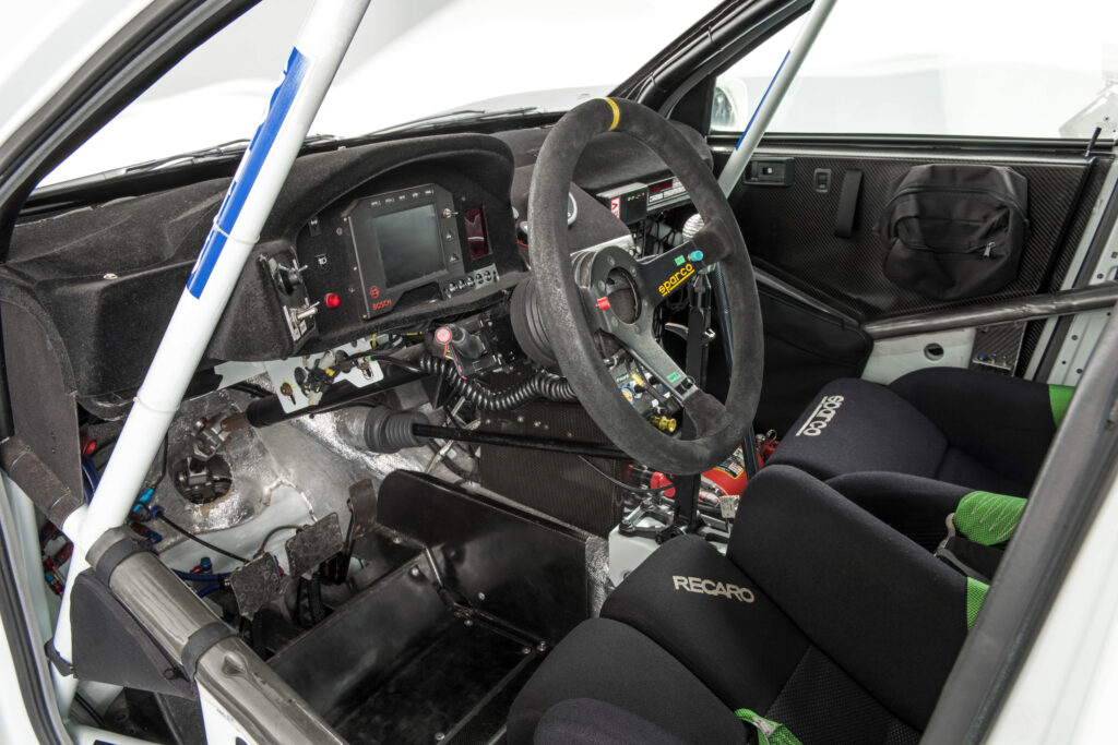 Race car cockpit interior with a Sparco steering wheel, Recaro racing seats, digital dashboard, and white roll cage.