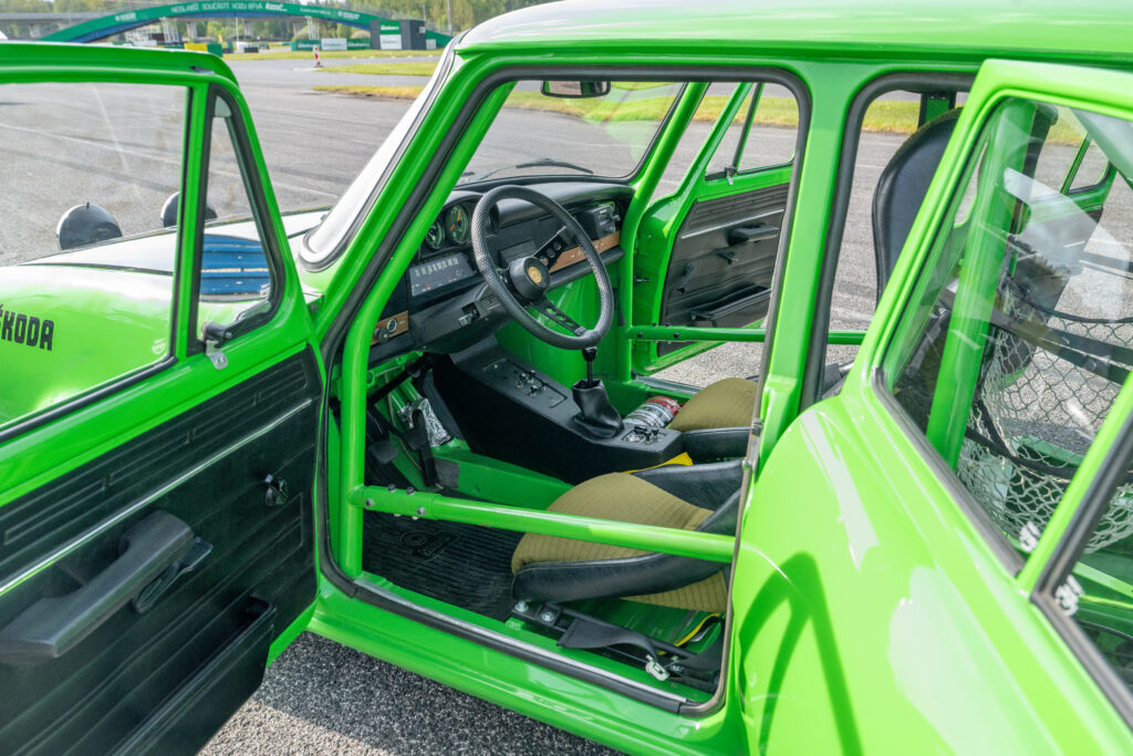 Interior of a vintage green Skoda rally car with a roll cage, racing bucket seats, and open doors on a racetrack.