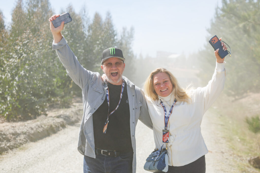 Excited man and woman celebrating with phones raised, wearing WRC all-access passes on a dusty rally road.