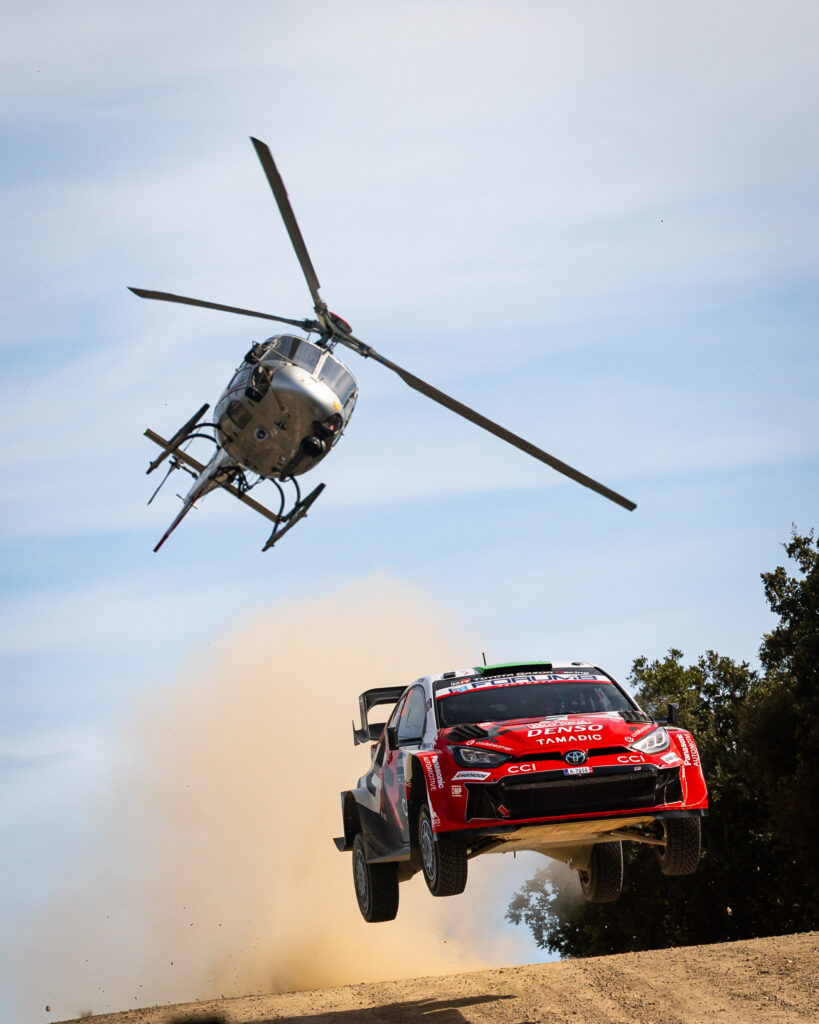 Toyota GR Yaris WRC rally car jumping mid-air on a dirt road with a tracking helicopter overhead.