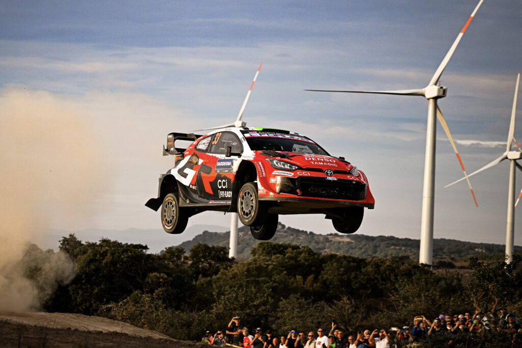 Toyota GR Yaris Rally1 car jumping high over a dirt crest with wind turbines in the background at Rally Sardegna.