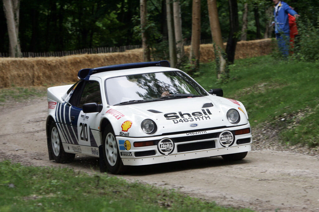 Classic white Ford RS200 Group B rally car with Shell branding driving on a dirt road course.