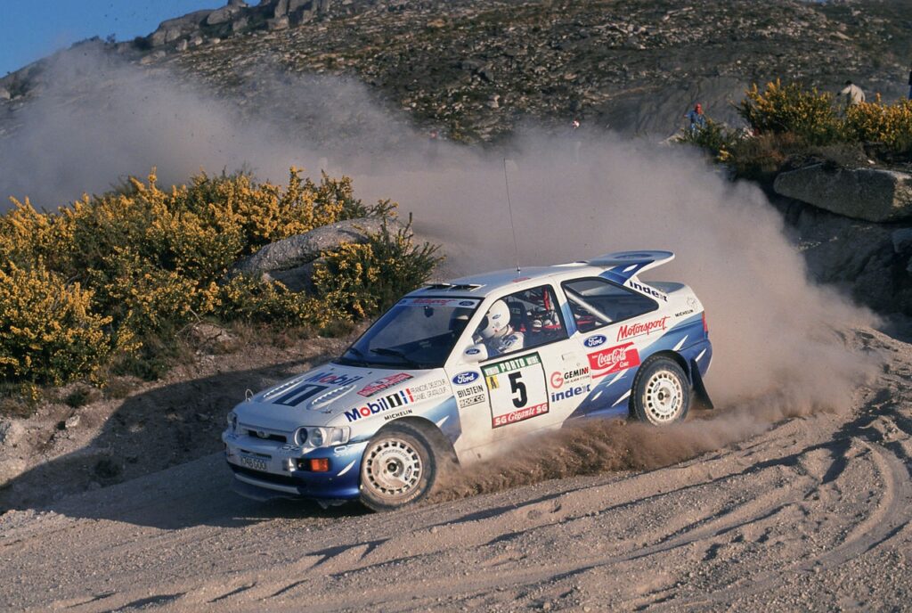 Ford Escort RS Cosworth rally car drifting on a dusty gravel road during a championship race.