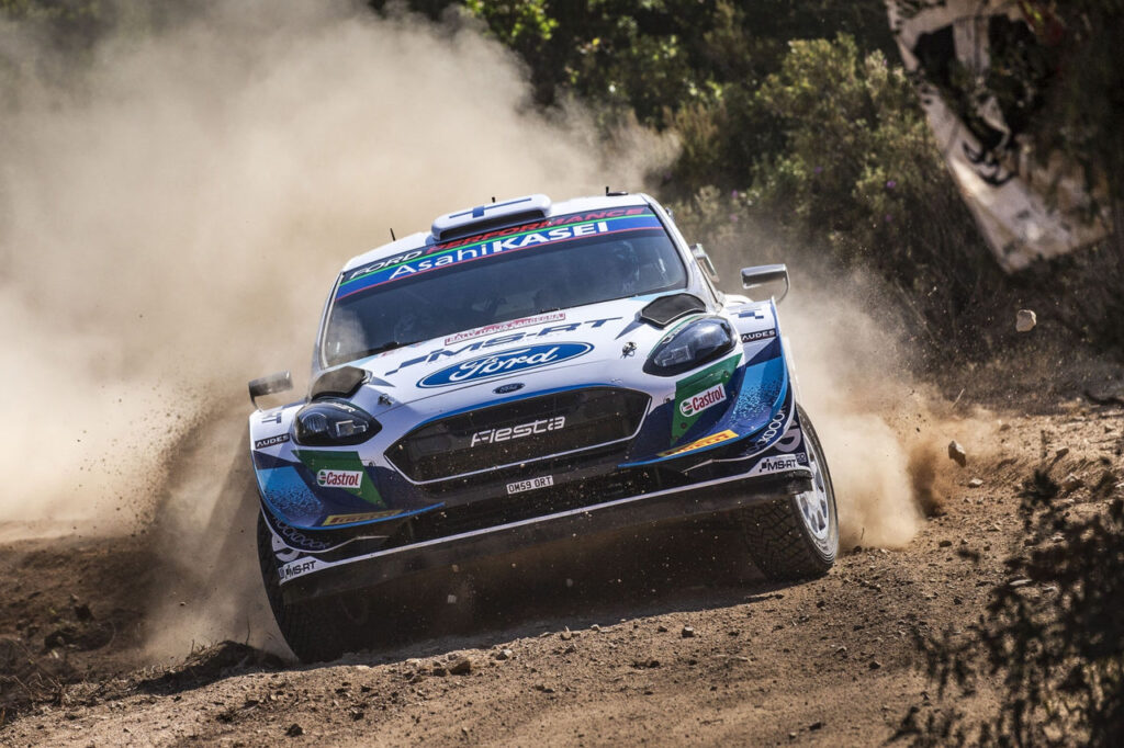 M-Sport Ford Fiesta WRC rally car drifting and kicking up a cloud of dust on a dirt race track.