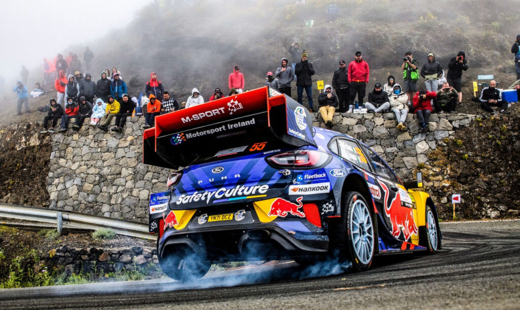 Ford Puma Rally1 car drifting with tire smoke on an asphalt mountain road during a WRC rally stage.