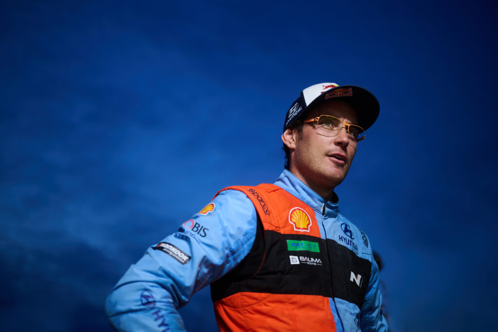 Thierry Neuville, WRC rally driver for Hyundai Motorsport, wearing a racing suit and cap against a blue sky.