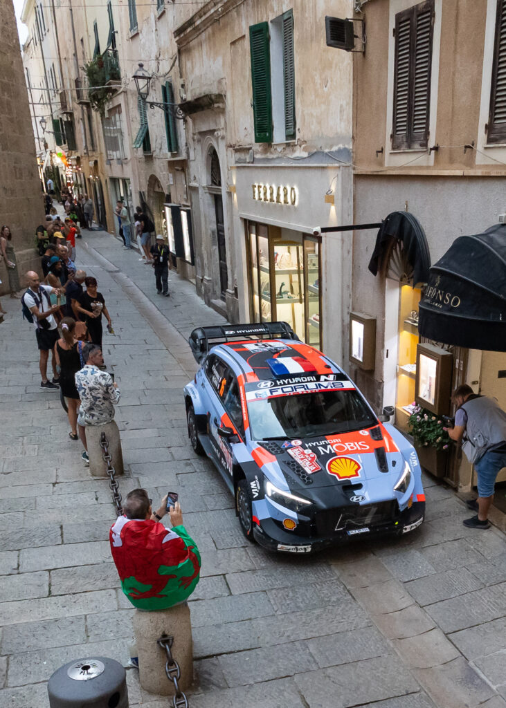 WRC Hyundai i20 N Rally1 car driving through a narrow Italian street during Rally Italia Sardegna with spectators.
