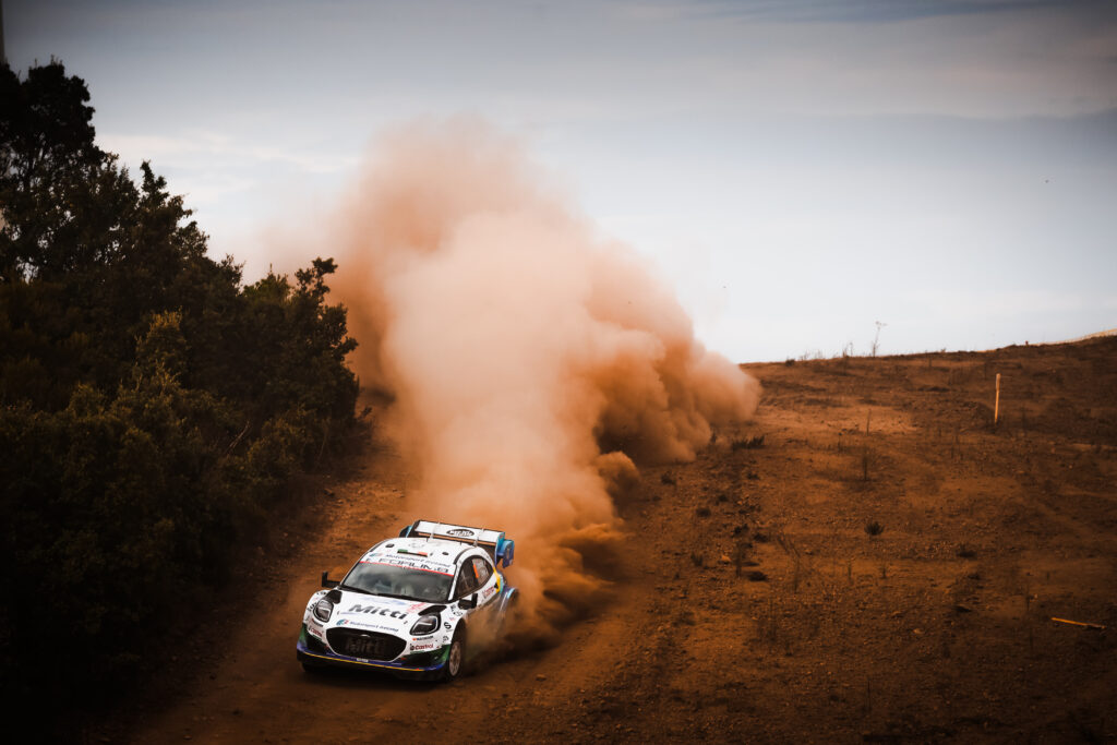 Rally car drifting on a dirt track, kicking up a massive dust cloud during an off-road motorsport race.
