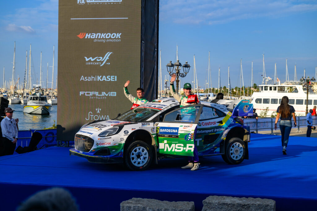 Jon Armstrong celebrates on the Rally Italia Sardegna podium next to his MS-RT Ford Fiesta Rally3 car.