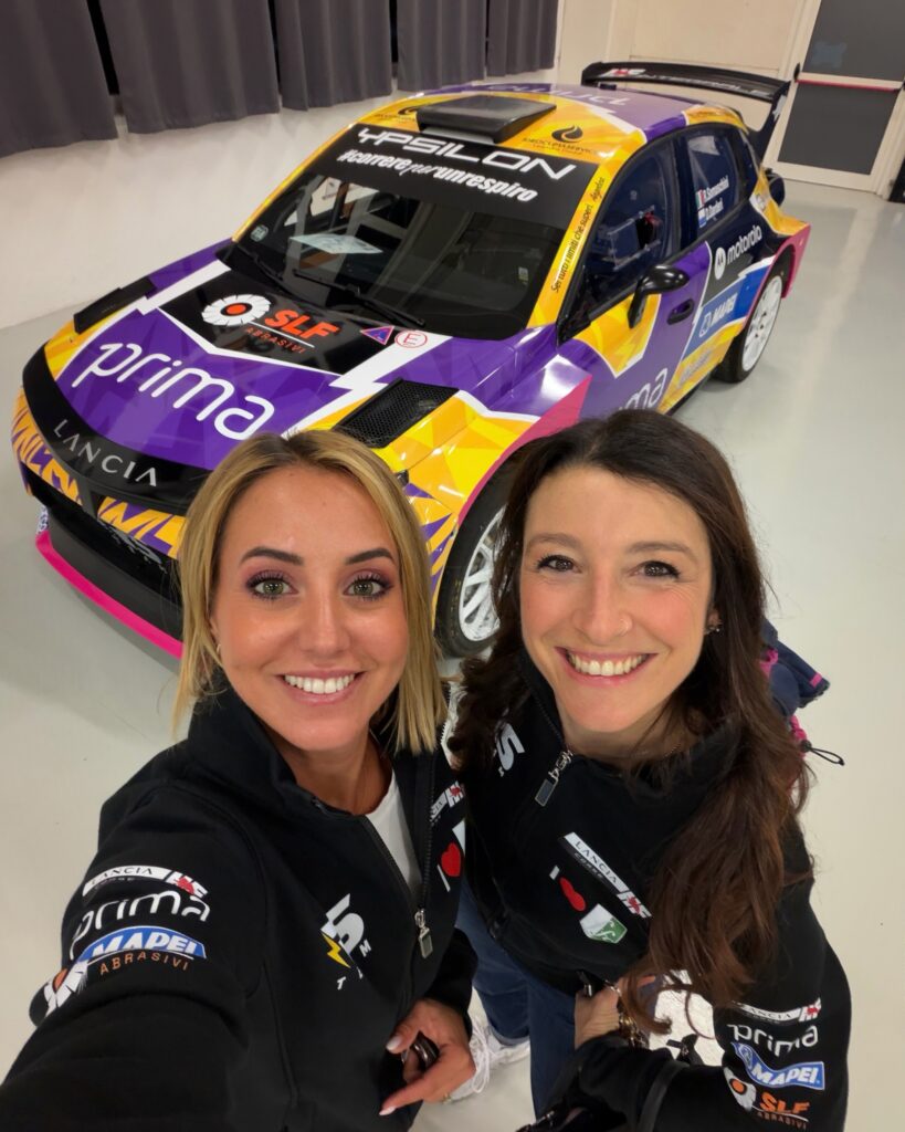Two women in Lancia Corse jackets take a selfie in front of a purple Lancia Ypsilon rally car.