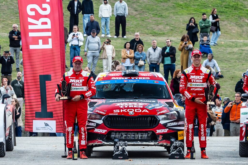 Two rally drivers in red race suits hold trophies beside a red MRF Tyres Skoda rally car with crowd in background.