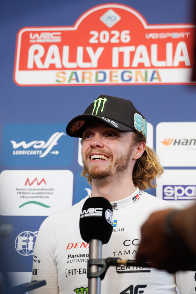 Oliver Solberg smiling at Rally Italia Sardegna 2026 interview, wearing WRC racing gear and a Monster Energy cap.