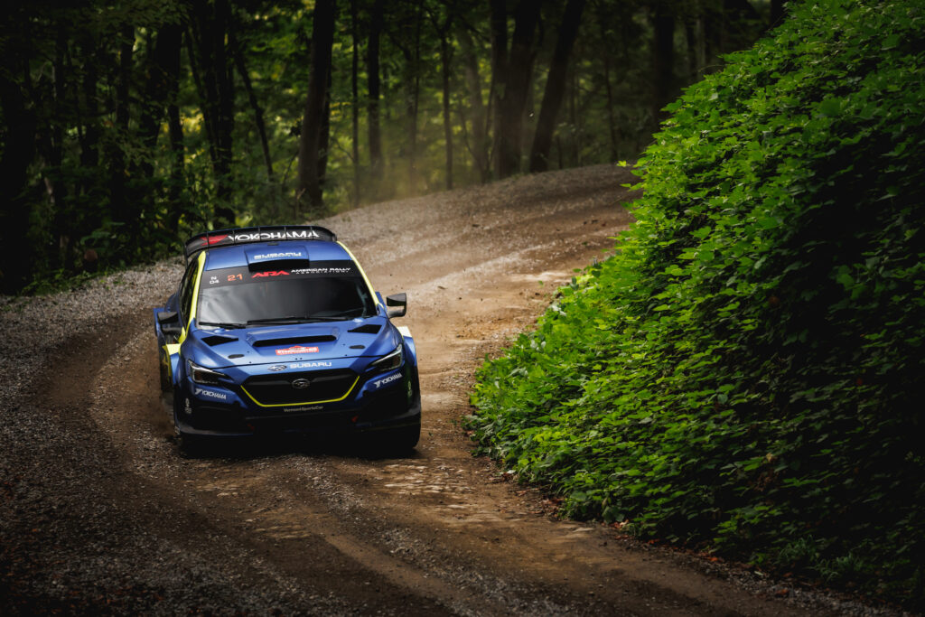 Blue Subaru WRX rally car driving fast around a curve on a dirt forest road during a rally race.